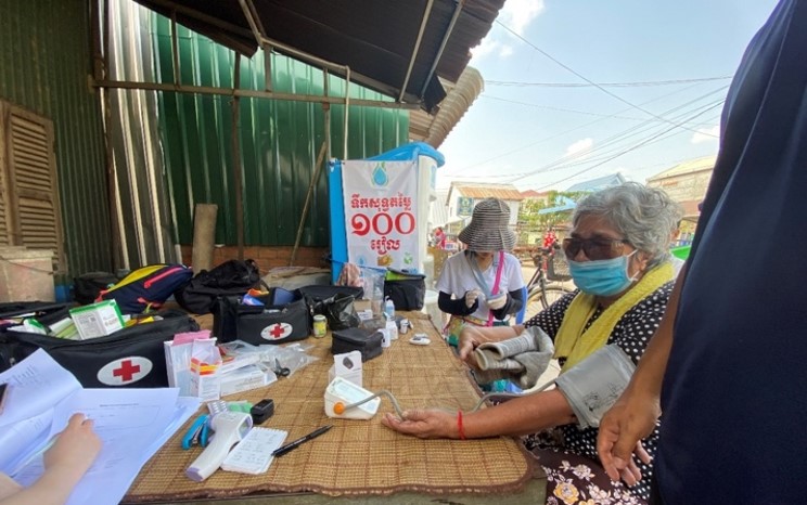 The image depicts a makeshift health check-up station outdoors in Phnom Penh, Cambodia. It's staffed by medical interns, as indicated by the presence of medical supplies and equipment on a table, such as blood pressure cuffs and a stethoscope. An elderly woman with gray hair, wearing a mask and a traditional patterned dress, is interacting with a medical intern. Other interns in the background are busy attending to patients or organizing supplies. The setting appears informal, with the focus on providing basic health services to the local community.
