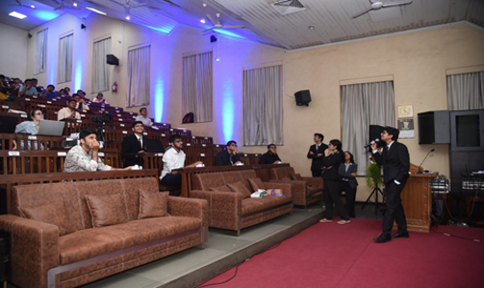 This image shows a formal indoor presentation or lecture setting. A speaker, dressed in a suit, stands at the front holding a microphone while addressing an audience. The audience, seated in tiered rows, includes individuals attentively listening and engaging with the speaker. The room features soft brown sofas in the front, wooden desks, and chairs for the audience, with blue ambient lighting and large windows covered by curtains. A podium and AV equipment are visible at the front, suggesting a professional or academic event."