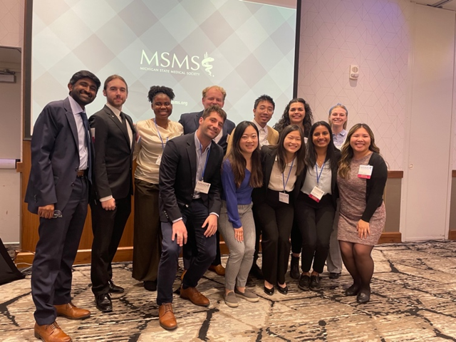 This image features a group of medical students and professionals posing together at the Michigan State Medical Society (MSMS) House of Delegates meeting. The attendees are dressed professionally and stand closely together, smiling at the camera in front of a presentation screen displaying the MSMS logo. The setting is a conference or meeting room, indicating a formal yet collegial atmosphere following an organized event or discussion.